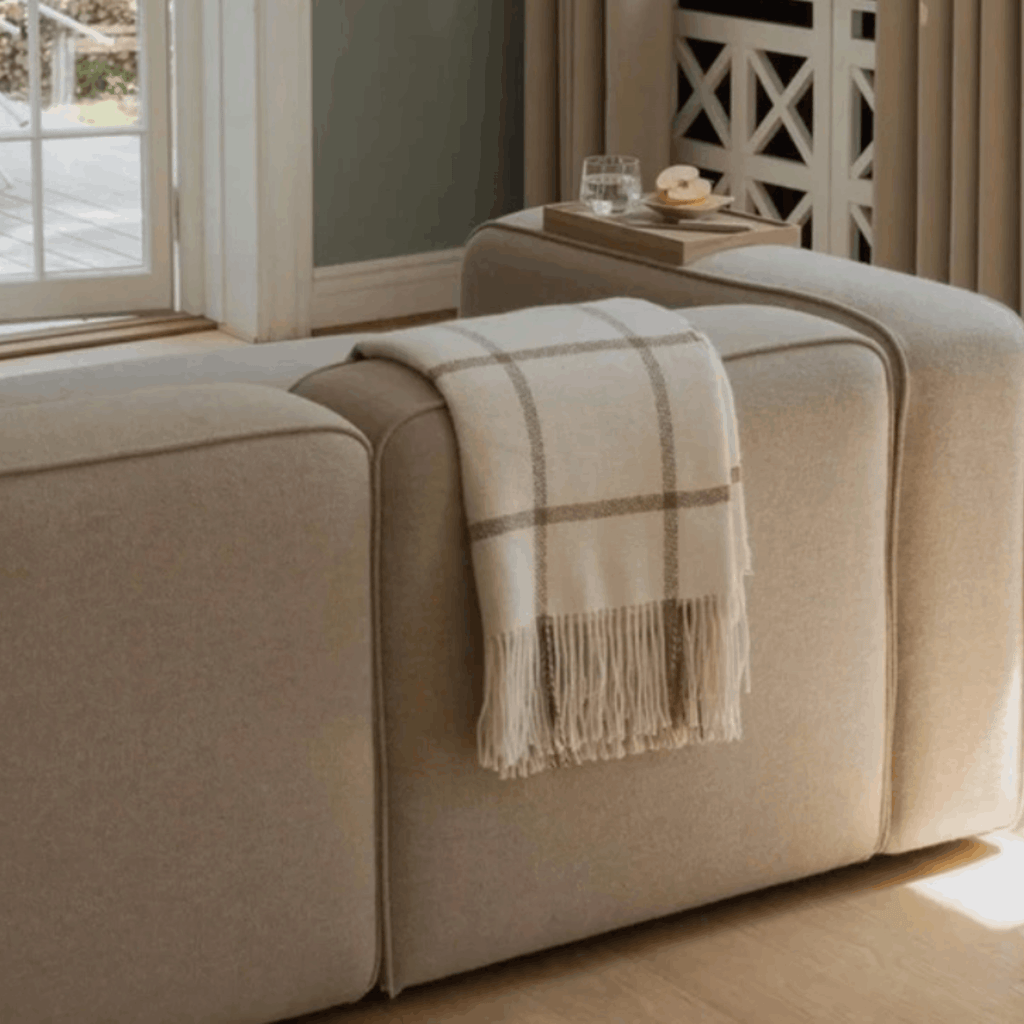 A beige upholstered sofa in a bright living room, with a cream plaid throw blanket draped over the seat. A small side table with a glass and tray sits beside the sofa near a window.