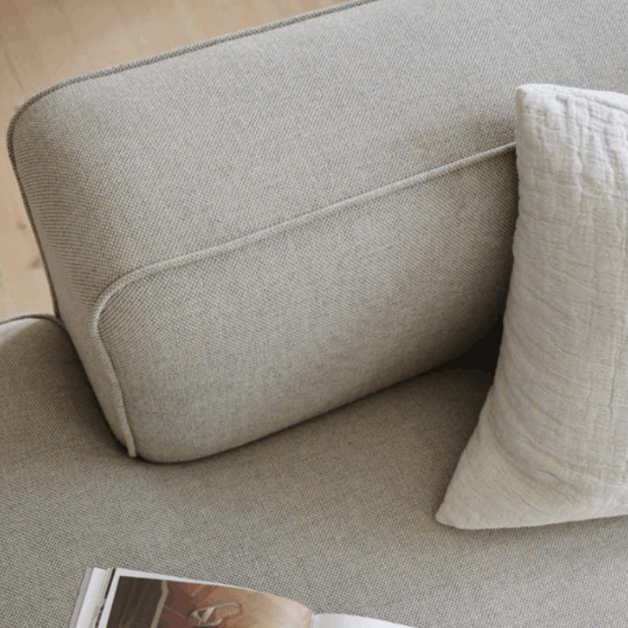 Close-up of a light beige upholstered sofa with a matching textured throw pillow and part of an open magazine visible in the foreground.