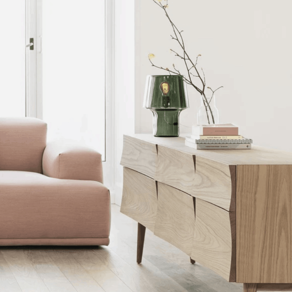 The Muuto Cosy Table Lamp by Harri Koskinen on a set of modern light oak wooden draws next to a pink sofa in a light modern living room.