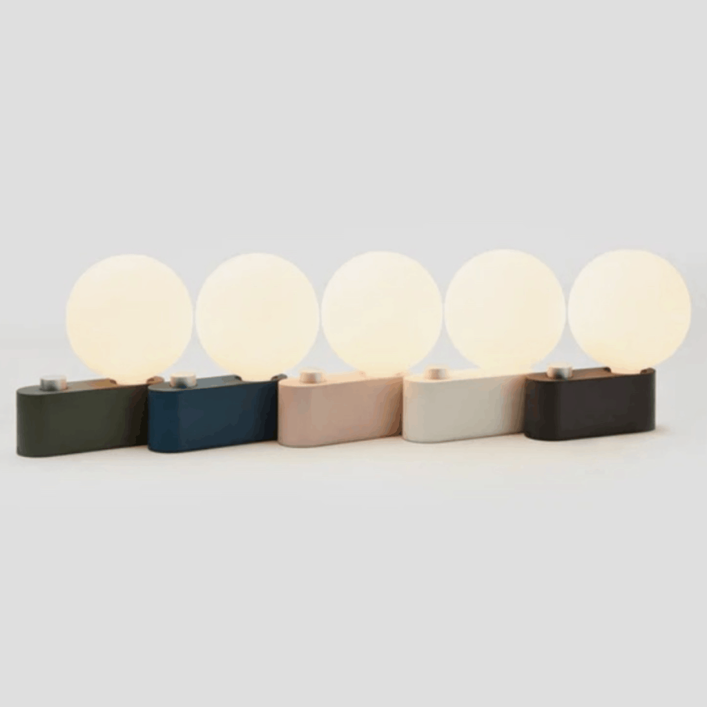 Five modern table lamps with rectangular bases in dark green, navy, blush pink, off-white, and black, each paired with a large spherical frosted shade glowing with warm light, arranged in a row against a light gray background.
