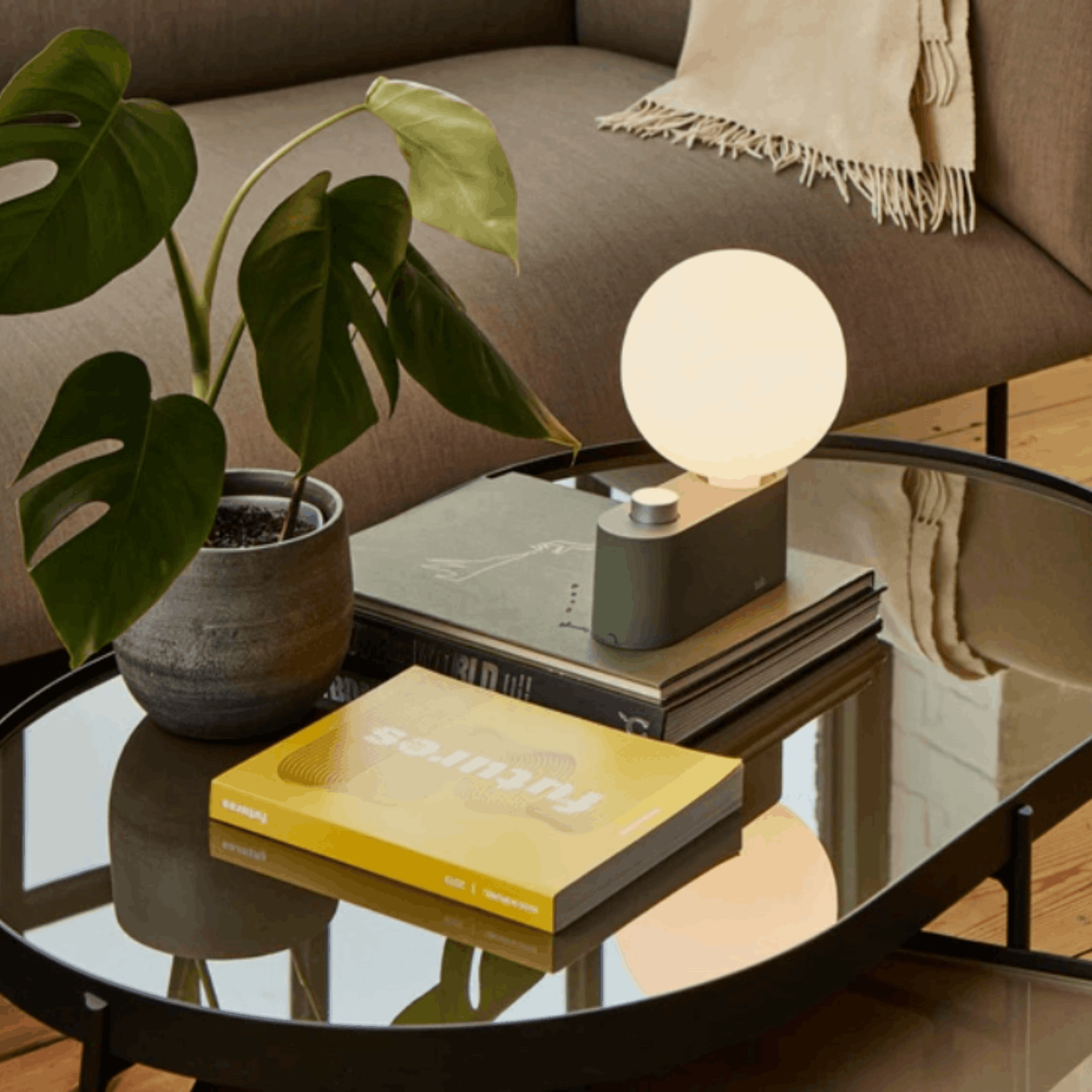 A modern spherical table lamp with a glowing white globe and a dark gray rectangular base sits on stacked books on a glass coffee table in a living room setting next to a potted monstera plant.