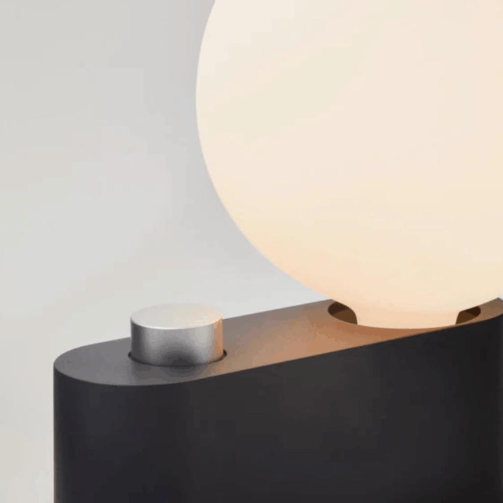 Close-up of a modern table lamp with a glowing warm white spherical shade and a matte black base featuring a brushed silver cylindrical accent.