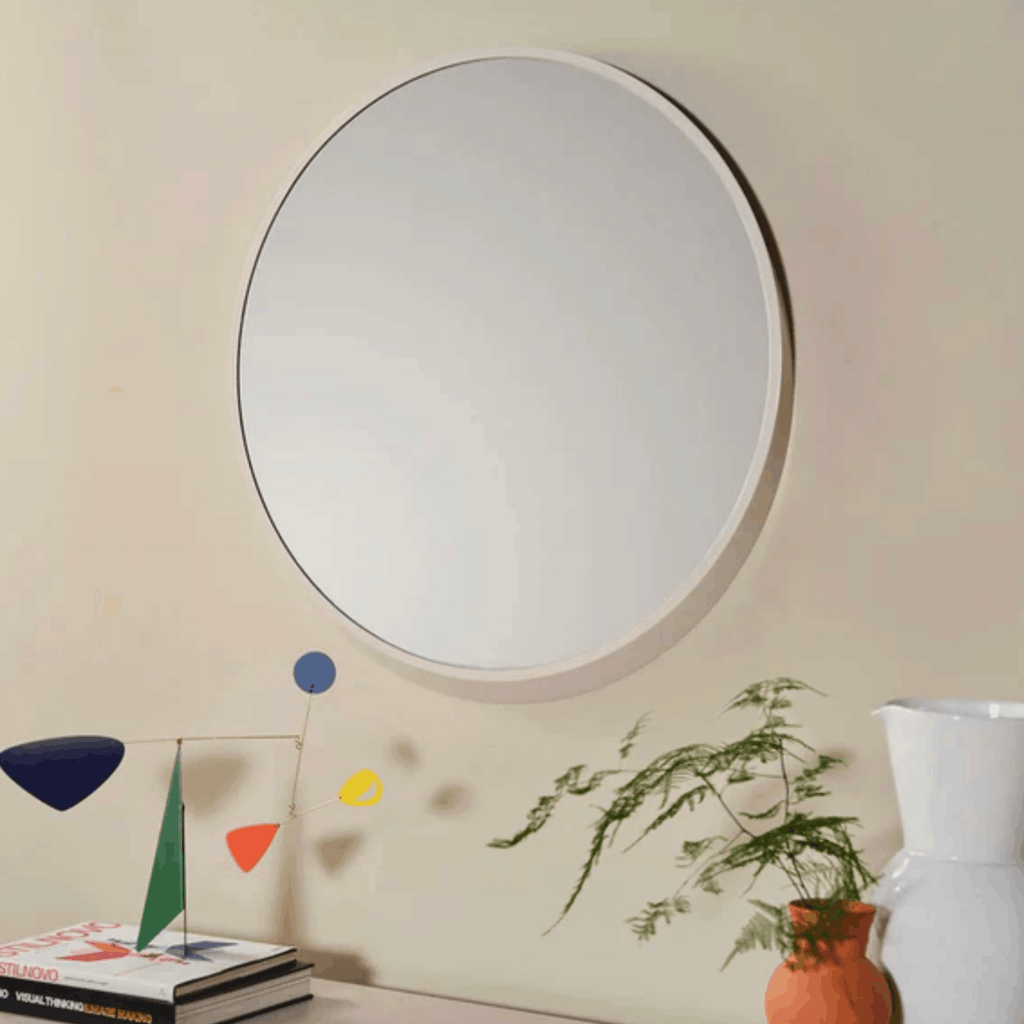 A large round wall mirror with a white frame hangs on a beige wall above a console table styled with books, a small colorful geometric sculpture, a potted fern, and a white vase.
