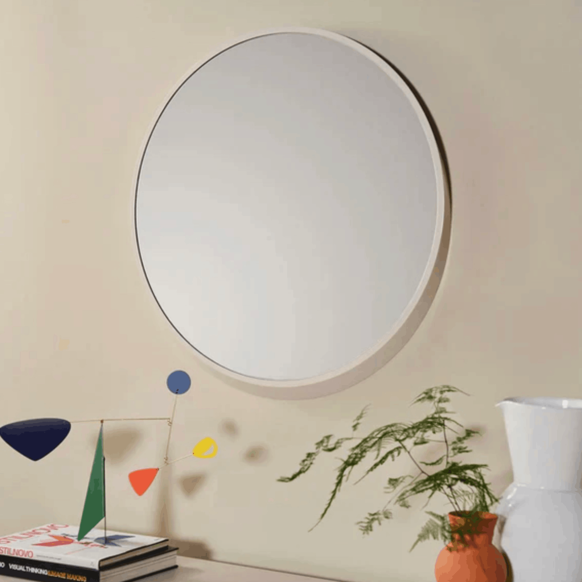 A large round wall mirror with a white frame hangs on a beige wall above a console table styled with books, a small colorful geometric sculpture, a potted fern, and a white vase.