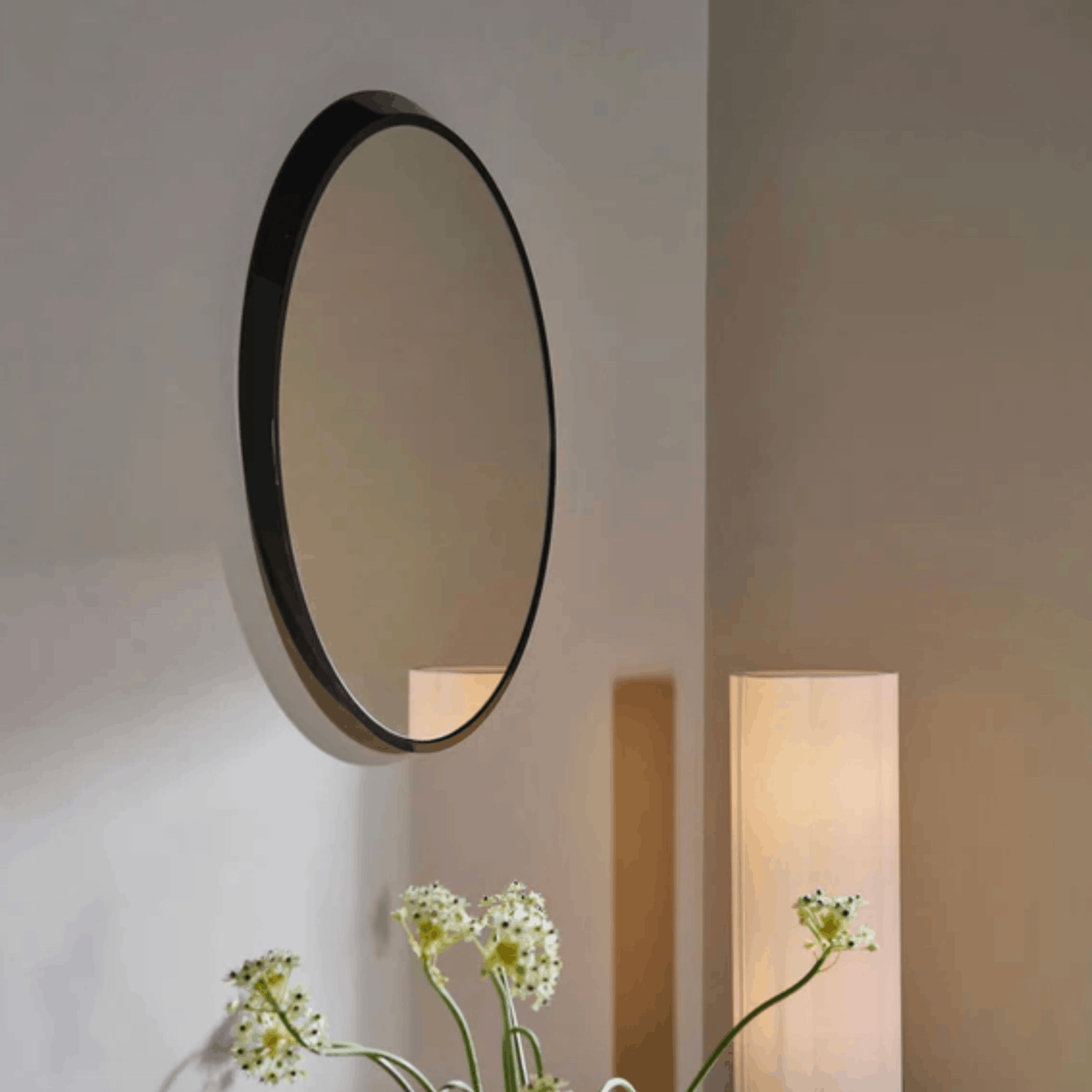 A round wall mirror with a thin black frame hangs on a beige wall in a softly lit interior. A floor lamp stands to the right, and white flowers are visible at the bottom left.
