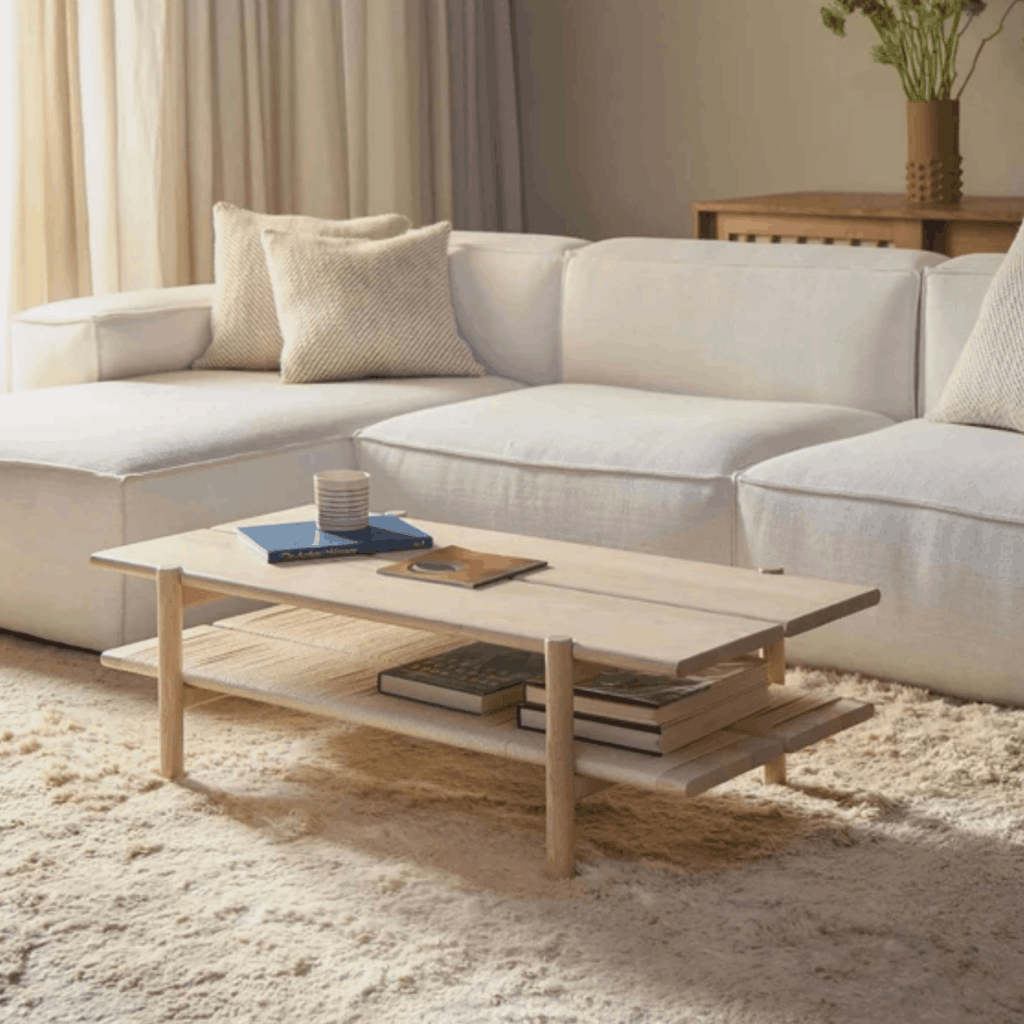 A light natural wood two-tier coffee table in a modern, neutral-toned living room setting with a white sectional sofa and beige rug.