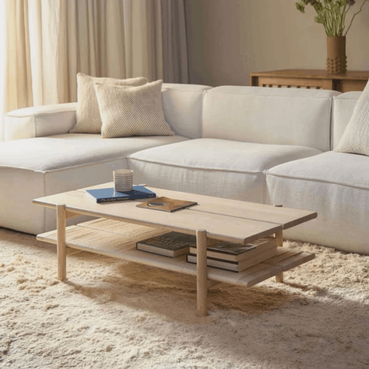 A light natural wood two-tier coffee table in a modern, neutral-toned living room setting with a white sectional sofa and beige rug.