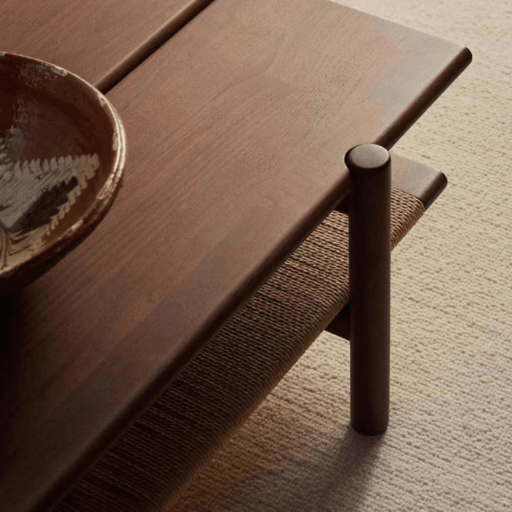 Close-up of a dark brown wooden table with a cylindrical leg and a woven seat cushion, shown in a styled interior setting with a ceramic bowl on the tabletop.