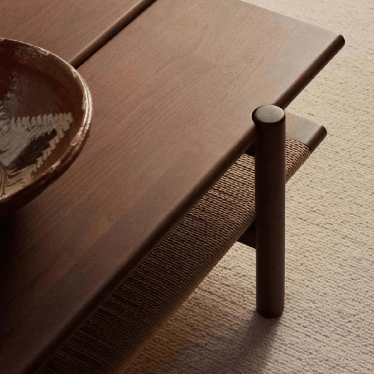 Close-up of a dark brown wooden table with a cylindrical leg and a woven seat cushion, shown in a styled interior setting with a ceramic bowl on the tabletop.