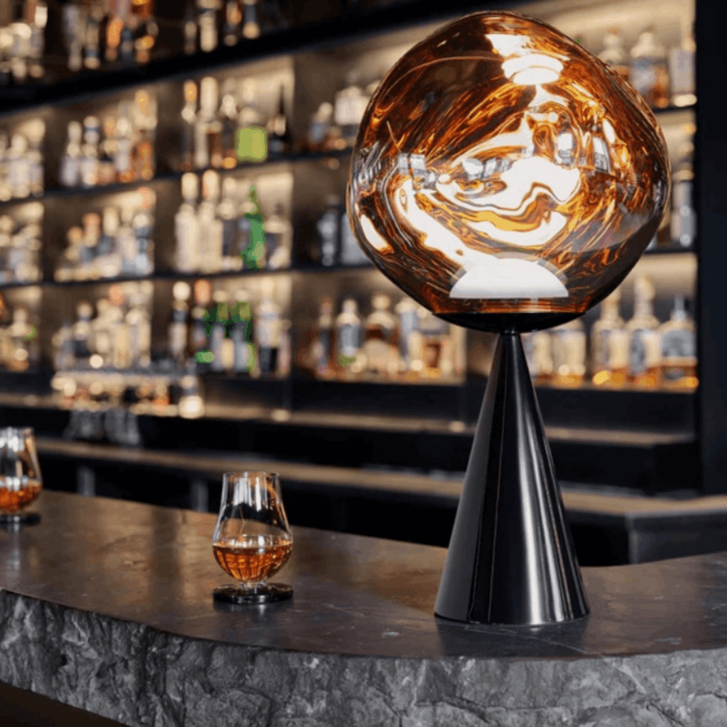 A modern table lamp with a large, glossy amber-brown spherical globe on a black conical base, styled on a dark stone bar counter in a bar setting with whiskey glasses and shelves of liquor bottles in the background.