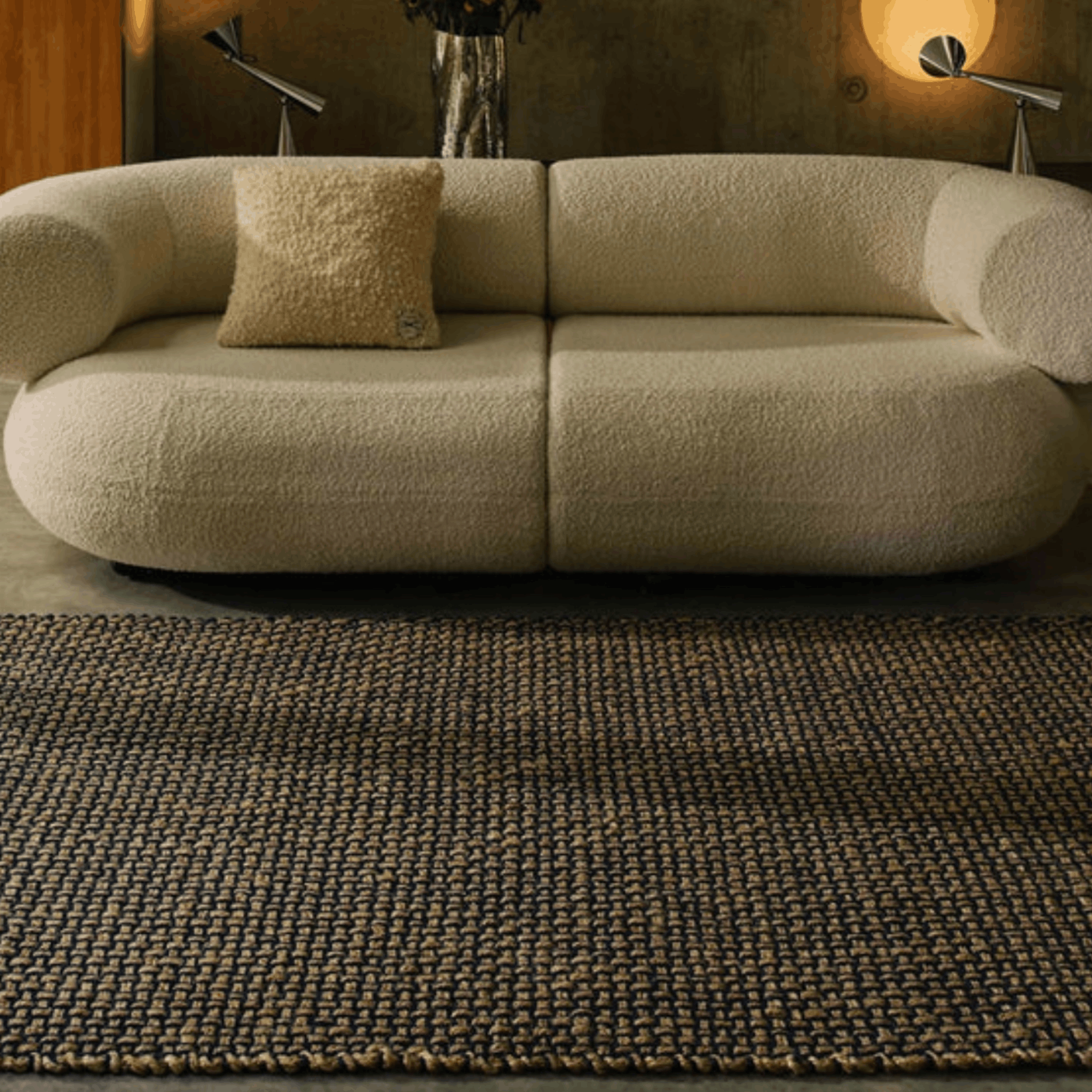 A cream bouclé sofa with a matching throw pillow in a styled living room, positioned on top of a Tome Dixon woven rug.
