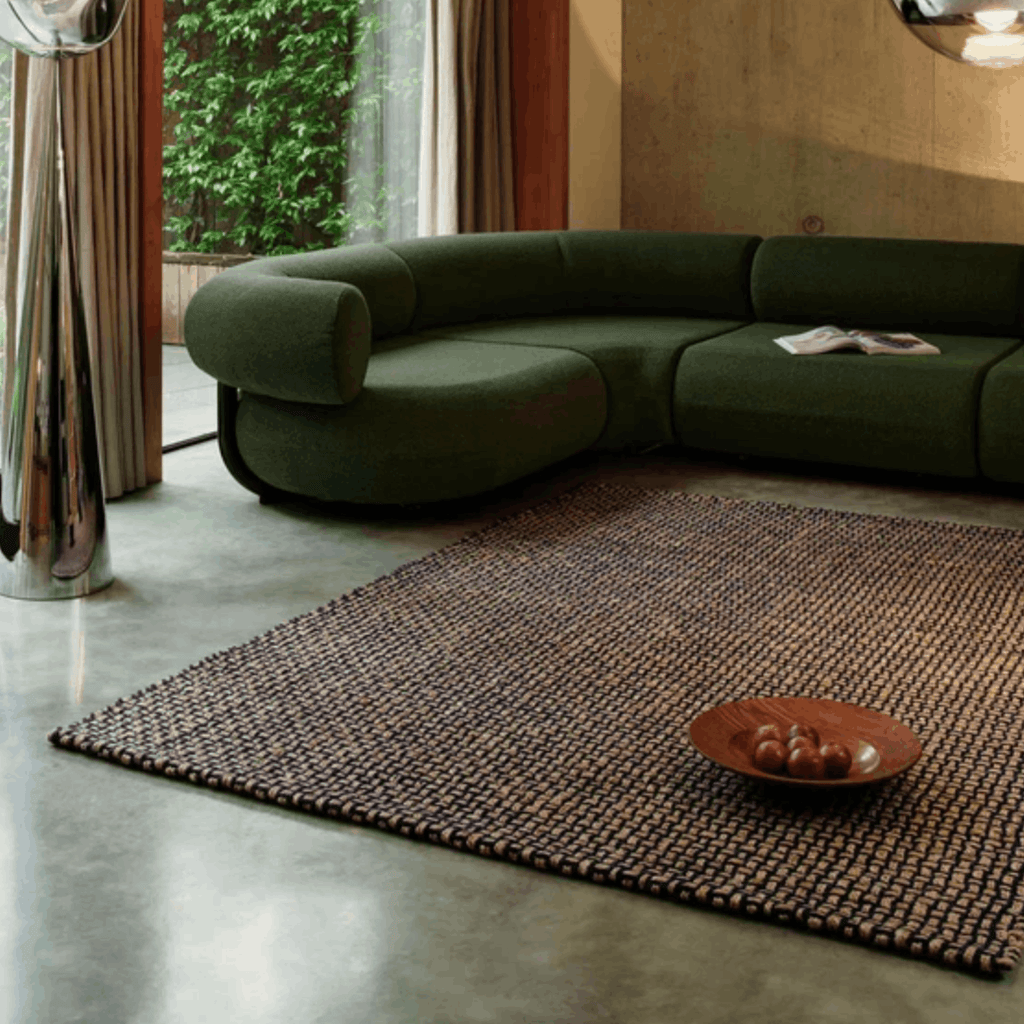 A modern curved sectional sofa upholstered in dark olive green fabric, placed in a contemporary living room with a large woven blue and beige Tom Dixon rug on a polished concrete floor.
