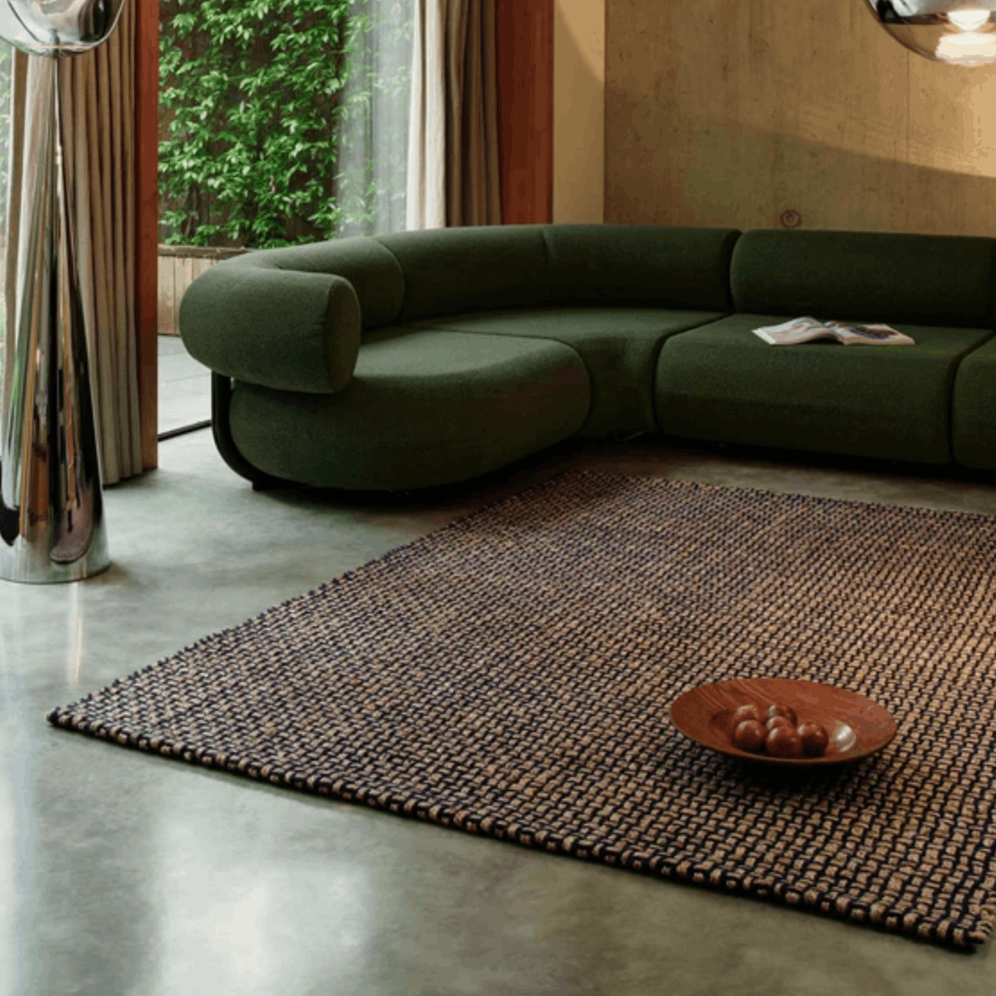 A modern curved sectional sofa upholstered in dark olive green fabric, placed in a contemporary living room with a large woven blue and beige Tom Dixon rug on a polished concrete floor.