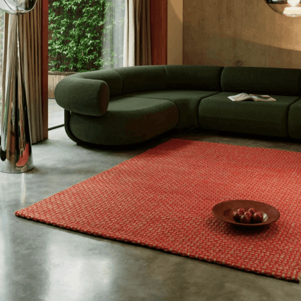 A modern living room scene featuring a prominent textured orange Tom Dixon rug on a polished concrete floor, with a dark green curved sectional sofa in the background.