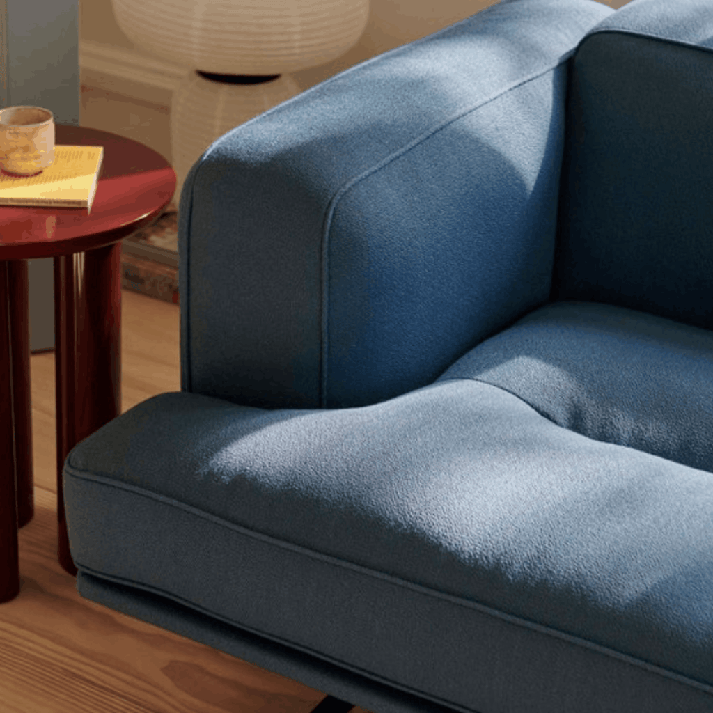 A close-up of a blue upholstered sofa in a real living-room setting, with a wooden side table, candle, and lamp visible in the background.