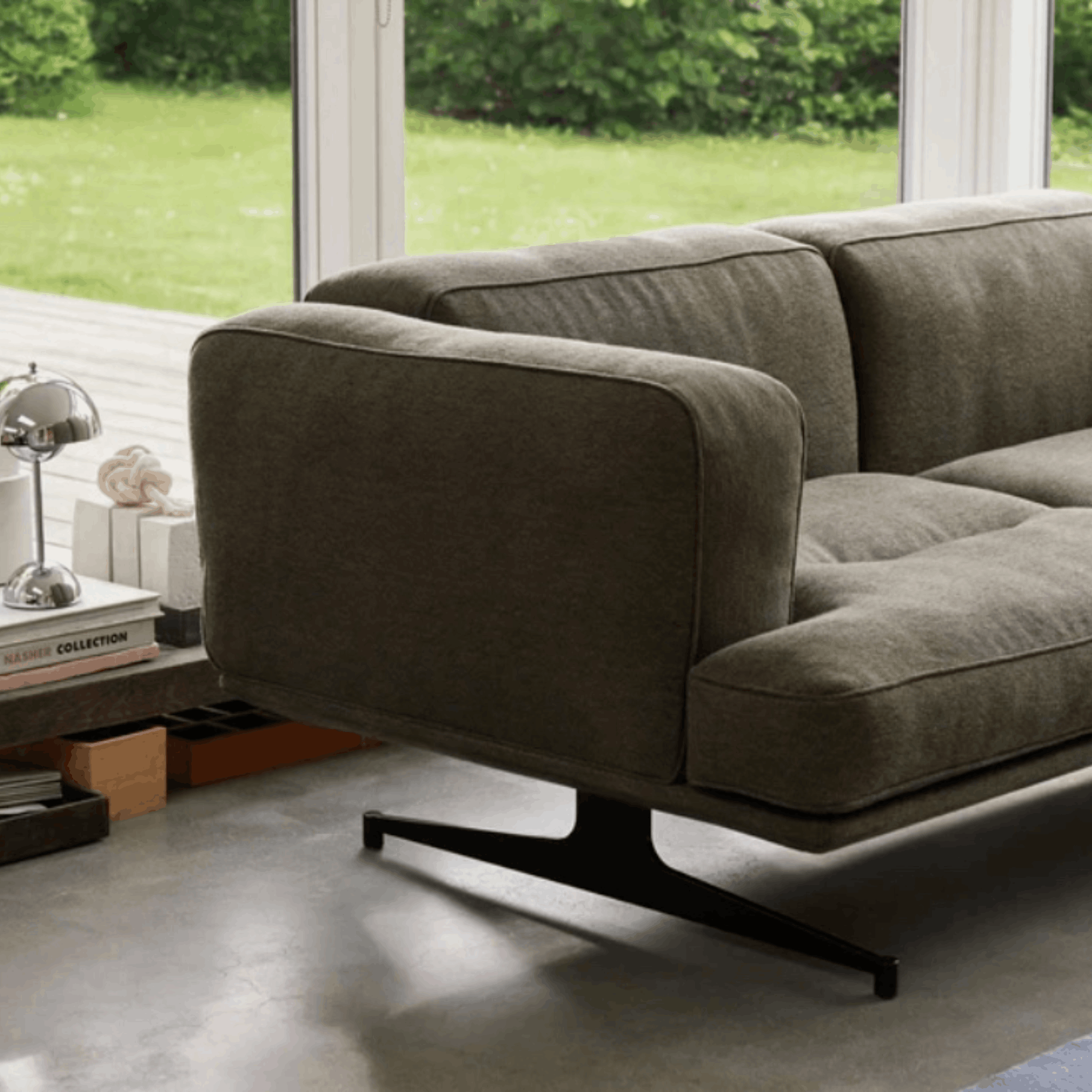A modern upholstered sofa in a muted taupe-gray fabric, shown in a real interior setting with a window view of green grass and trees, a side table with a lamp and books, and a concrete floor.