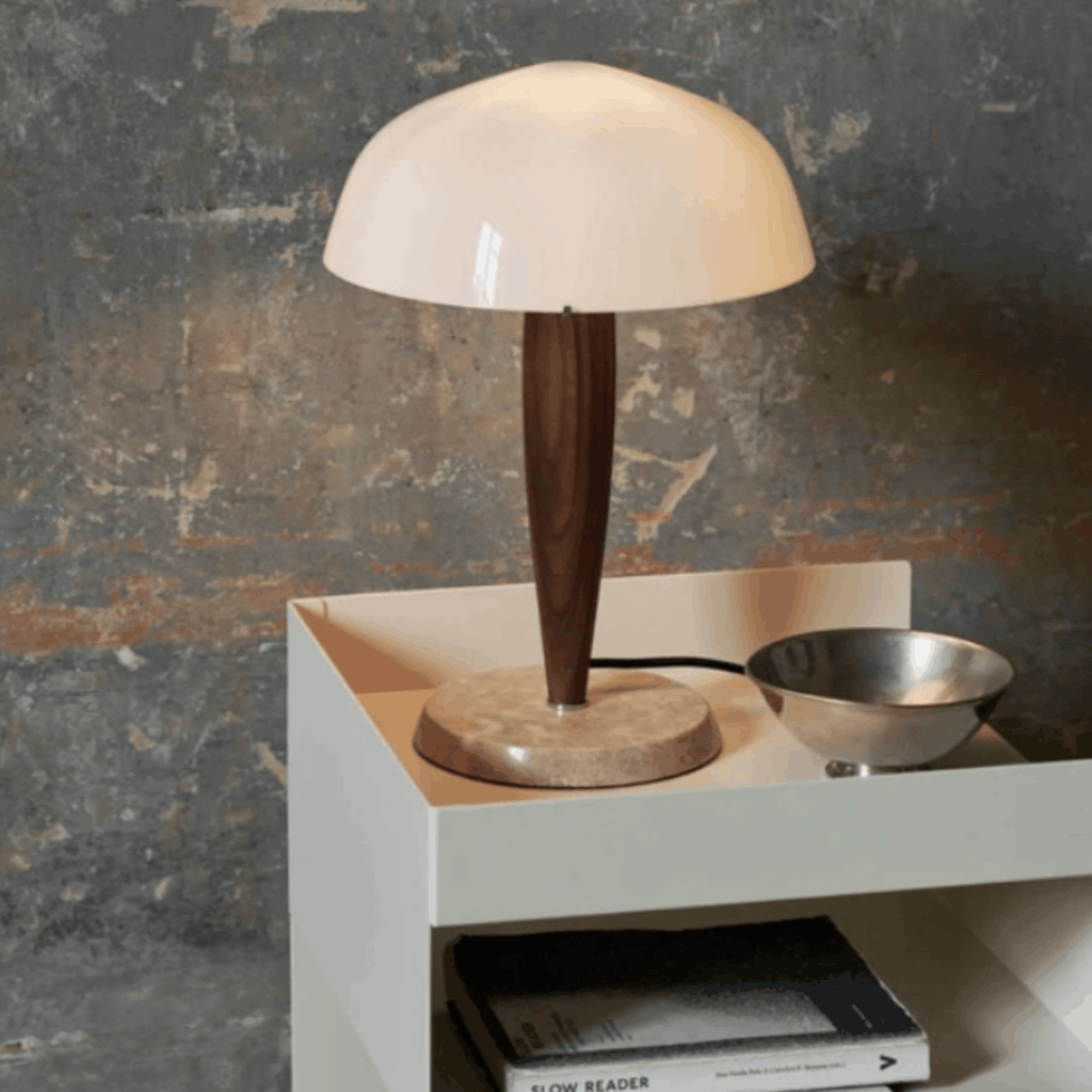 A mushroom-shaped table lamp with a glossy cream dome shade, a dark brown tapered wooden stem, and a round stone base, styled on a white side table next to a metal bowl against a textured gray wall.