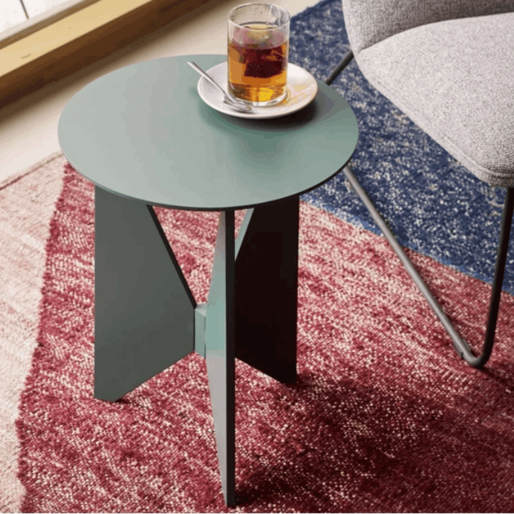 Treku Hiru Coffee Table by Estudi Manel Molina, featuring a compact round top and distinctive three-legged base in a coffee shop setting on a coloured rug with grey chair beside it.