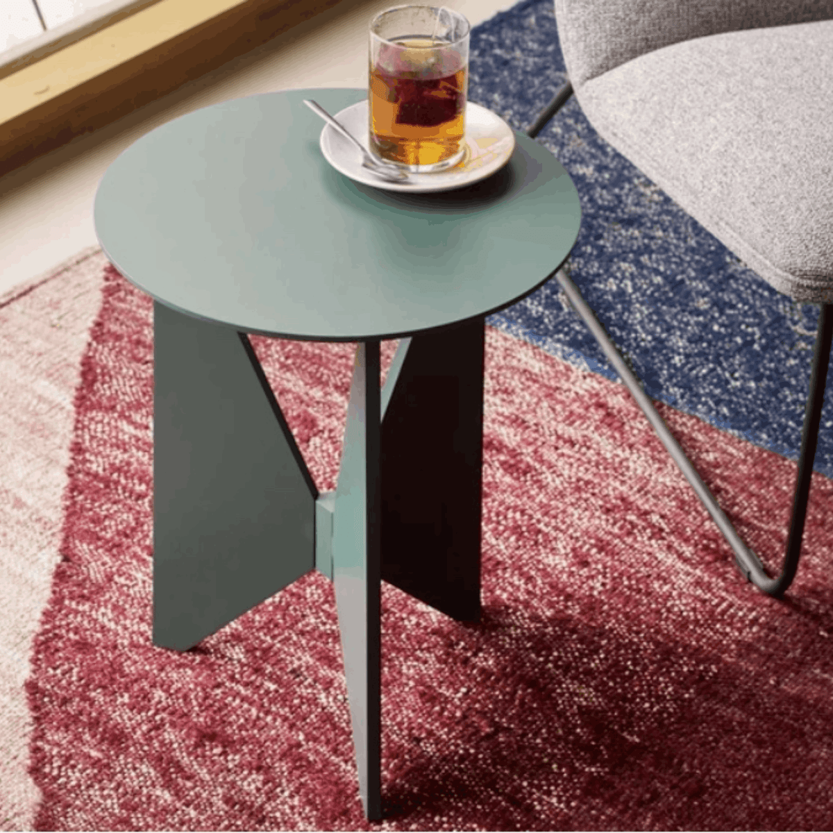 Treku Hiru Coffee Table by Estudi Manel Molina, featuring a compact round top and distinctive three-legged base in a coffee shop setting on a coloured rug with grey chair beside it.
