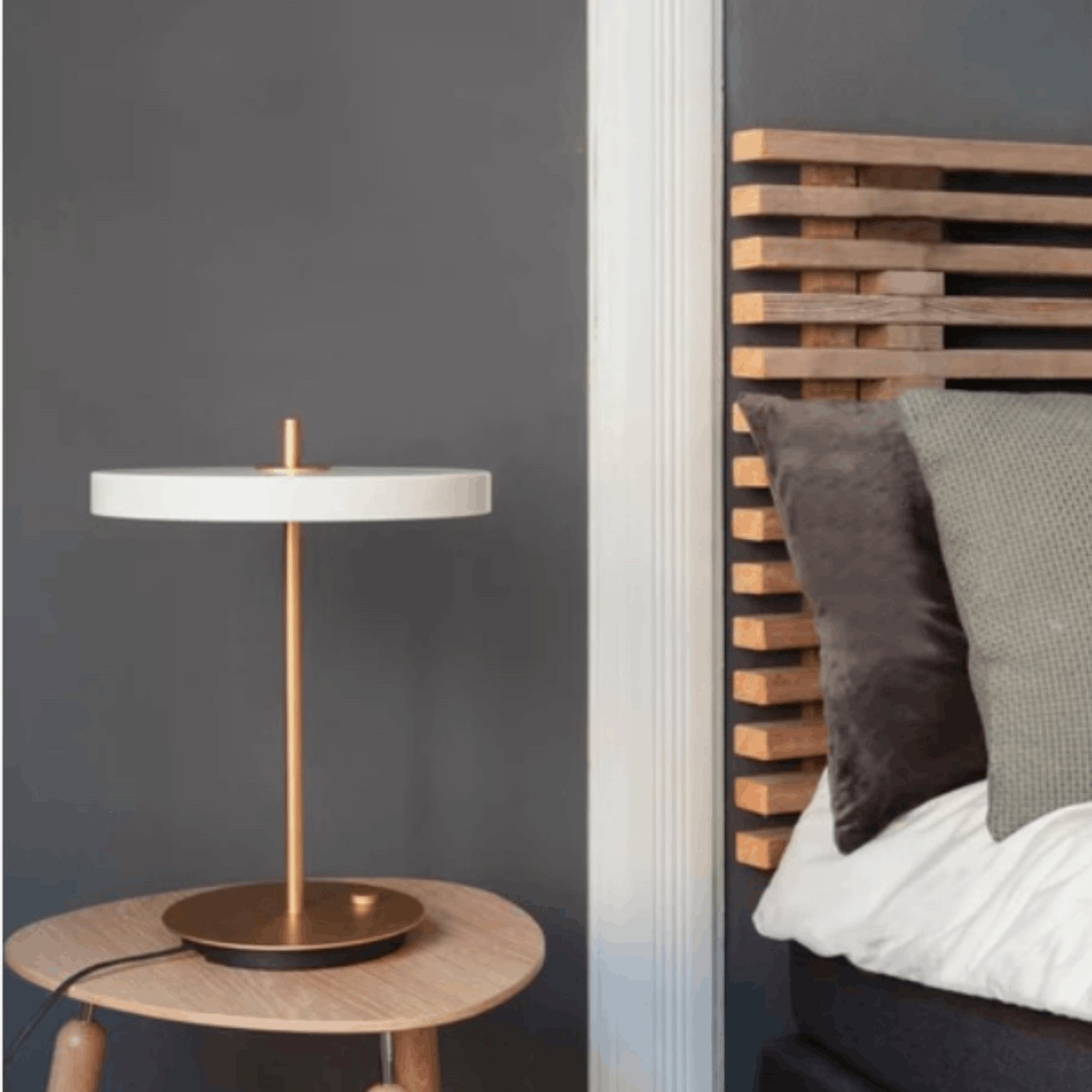 Modern table lamp on a round wooden side table next to a bed with wooden headboard and pillows.