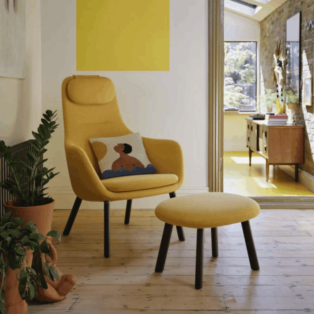 Yellow armchair with a cushion and matching ottoman in a room with plants and a window.