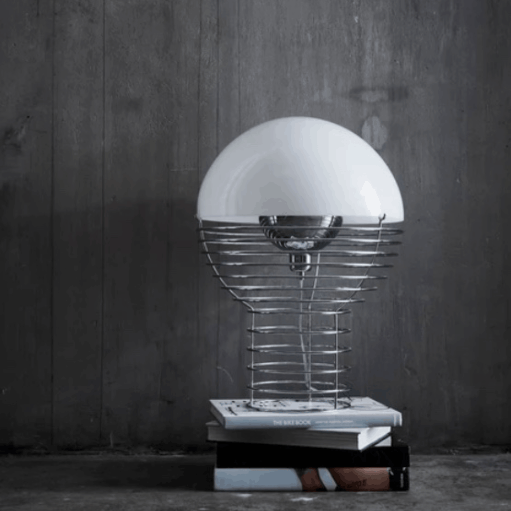 A modern table lamp with a large white dome shade, a silver wire spiral base, and a stack of books on a dark gray wood-paneled floor.