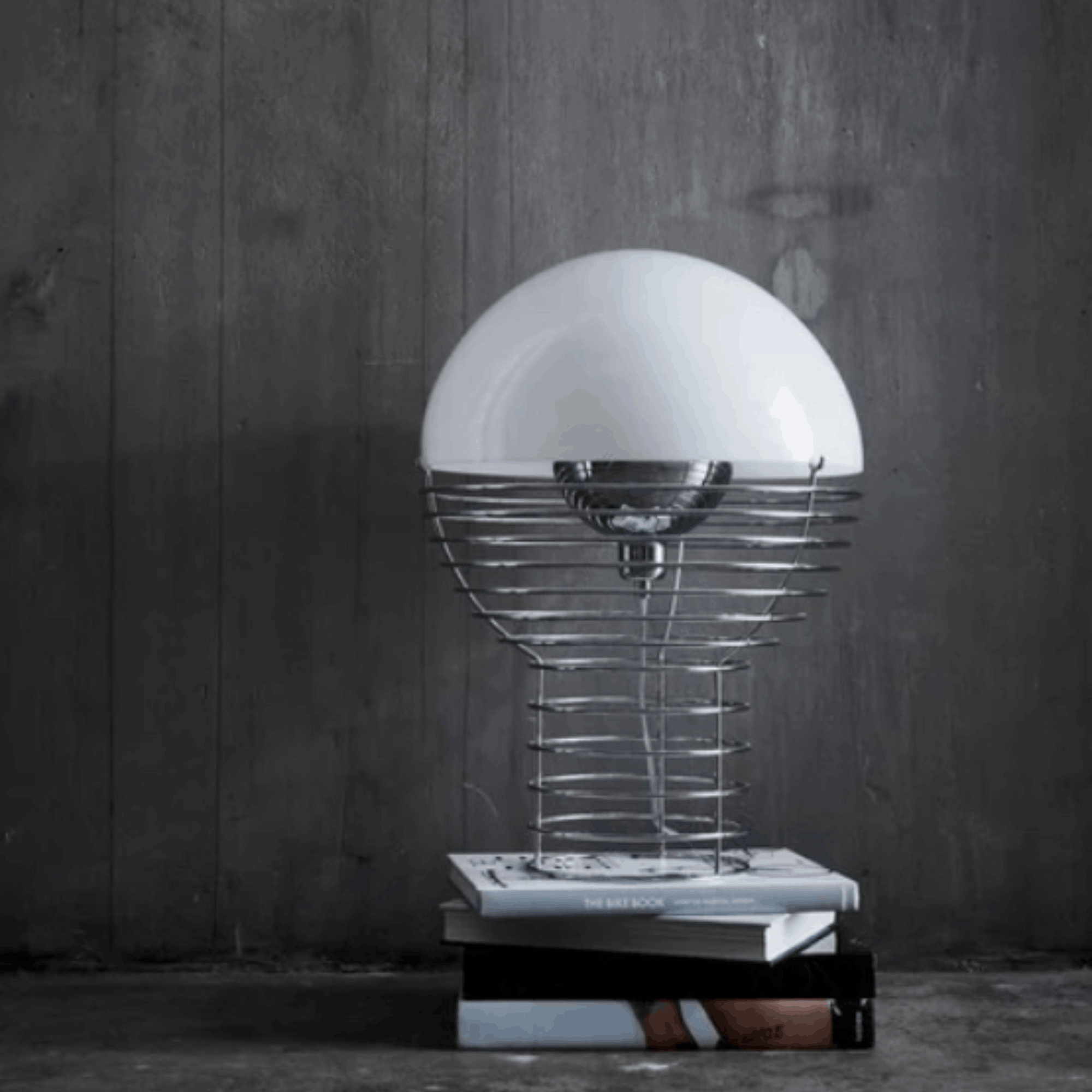 A modern table lamp with a large white dome shade, a silver wire spiral base, and a stack of books on a dark gray wood-paneled floor.